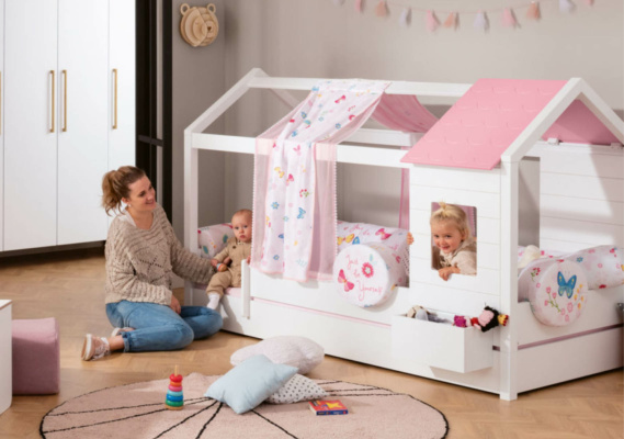 Kinderzimmer für zwei: Ein Spielbett in Form eines kleinen Hauses, dekoriert mit Schmetterlings- und Blumenmotiven in Rosa und Weiß. Eine Frau sitzt mit einem Baby auf dem Boden, während ein weiteres Kind fröhlich aus einem Fenster des Betts schaut. Der Raum ist kindgerecht eingerichtet mit weichen Kissen und Spielzeug.