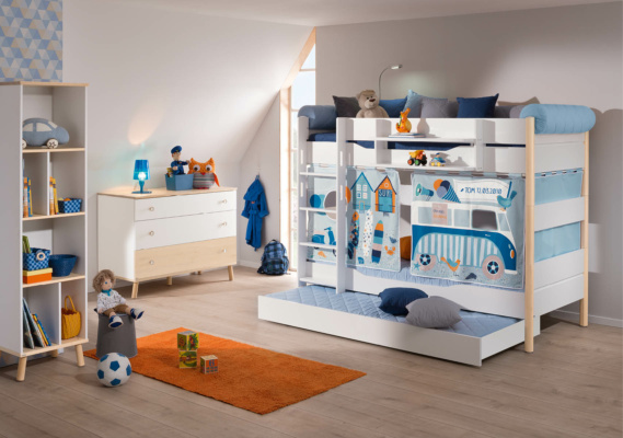 Geschwisterzimmer einrichten: Ein Kinderzimmer mit einem weißen Etagenbett, dekoriert mit bunten Stoffbahnen in Blau und Orange, die ein Strand- und Surfmotiv zeigen. Im Raum steht eine kleine Kommode, Regale mit Spielzeug und ein orangefarbener Teppich. Auf dem Boden liegen eine Puppe und eine Spielzeugkiste.