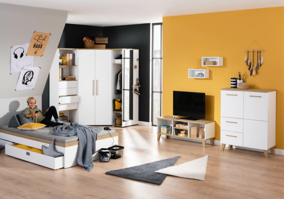 Jugendzimmer einrichten: Jugendzimmer mit modernen Möbeln in Weiß und Grau, kombiniert mit einer gelben Wand. Ein Junge sitzt auf einem Bett und schaut auf sein Handy. Ein Kleiderschrank, ein Fernseher, Regale und Dekorationen wie Traumfänger und Poster ergänzen den Raum.